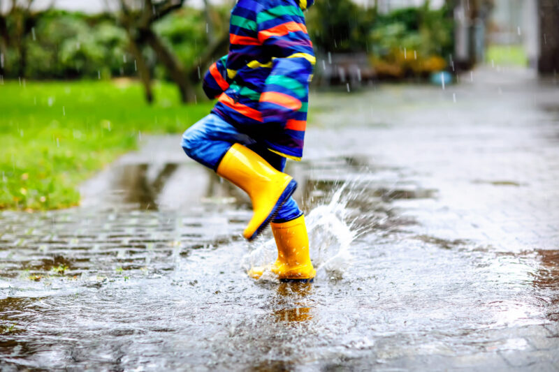 Close-up of child wearing yellow rain boots and splashing in a puddle. [Image © Adobe Stock]